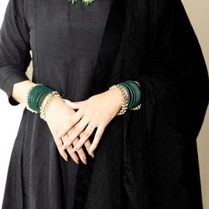 以下が含まれることがあります： 黒いドレスを着た女性が、緑と金のネックレスとそれに合わせた緑のバングルをつけている
