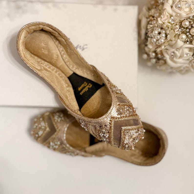 May include: A pair of gold-colored, embellished flats. The shoes feature intricate beadwork and a mesh overlay on the toe area. The soles are a darker shade of gold, and the interior is black. The shoes are placed on a white surface.