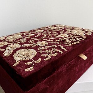 May include: A burgundy velvet box with intricate gold floral embroidery on the lid. The box has a rectangular shape and a small, gold-colored ribbon pull. The embroidery features detailed flowers and leaves.