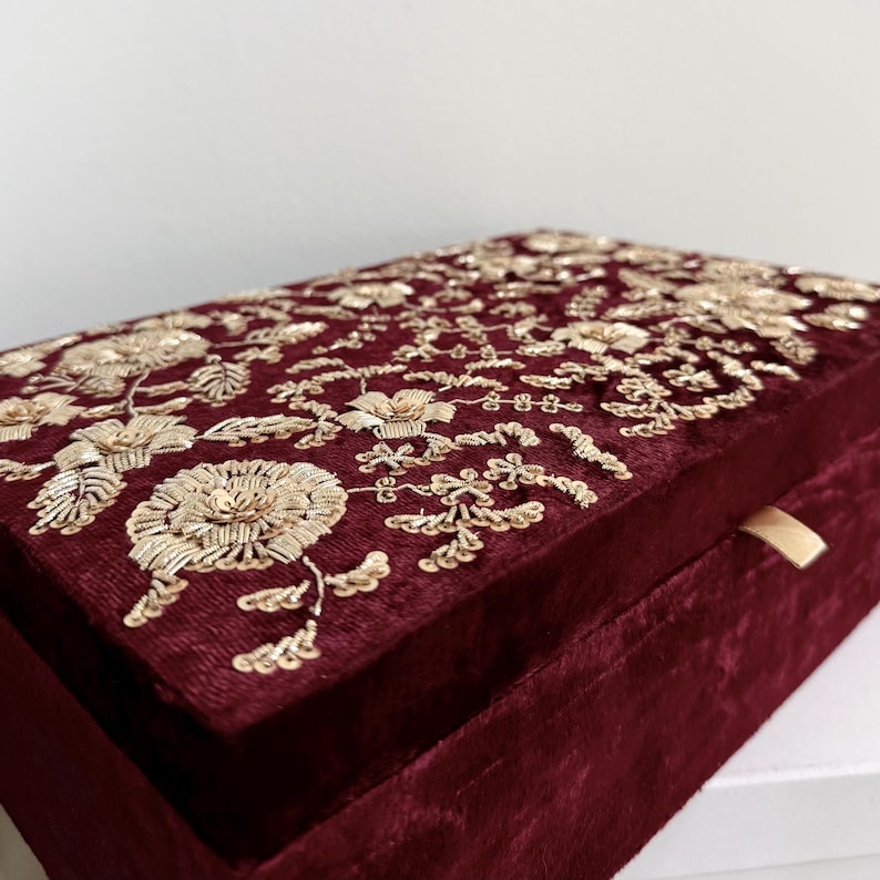 May include: A burgundy velvet box with intricate gold floral embroidery on the lid. The box has a rectangular shape and a small, gold-colored ribbon pull. The embroidery features detailed flowers and leaves.