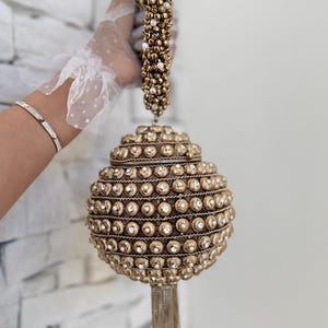 May include: A gold-colored, beaded handbag with a round shape and a tassel. The handle is also adorned with beads and pearls. The bag is held up by a person wearing a white lace glove and a silver bracelet.