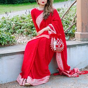 May include: A woman wearing a red saree with silver embroidery and a red beaded bag. The saree has a sparkly texture.