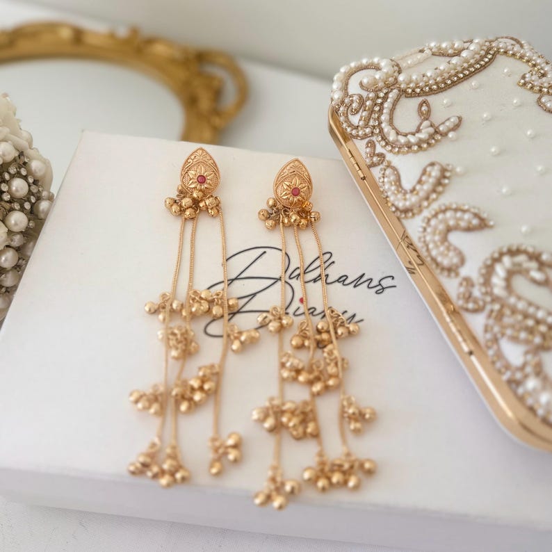 May include: Gold-toned dangle earrings with a teardrop-shaped top, a small red gem, and multiple dangling chains with small ball accents. The earrings rest on a white box with the text "Dalhans Diaries". A decorative clutch with pearl and gold accents is also visible.