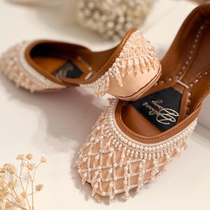 May include: A pair of beige leather shoes with intricate beaded embellishments. The shoes have a pointed toe and a low heel. The beads are arranged in a geometric pattern and create a delicate fringe effect.