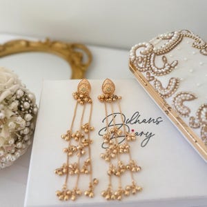 May include: Gold-toned dangle earrings with intricate detailing. Each earring features a decorative top with a small gemstone and multiple strands of small, dangling beads. The earrings rest on a white box with the text "Dulhans Diary".