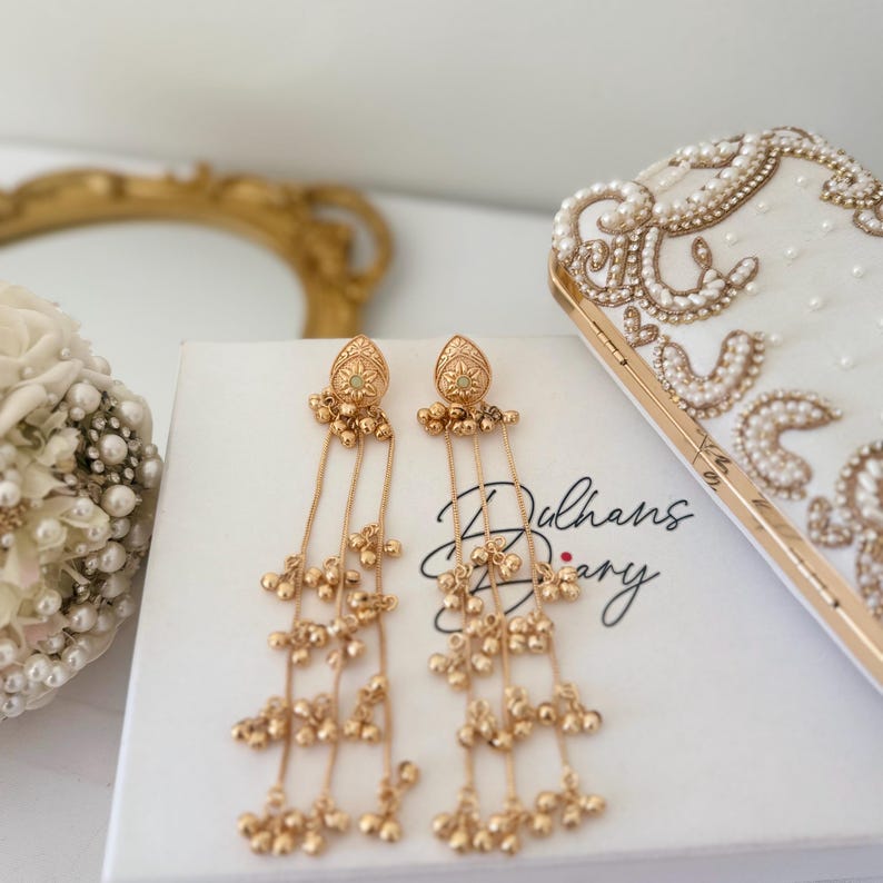 May include: Gold-toned dangle earrings with intricate detailing. Each earring features a decorative top with a small gemstone and multiple strands of small, dangling beads. The earrings rest on a white box with the text "Dulhans Diary".
