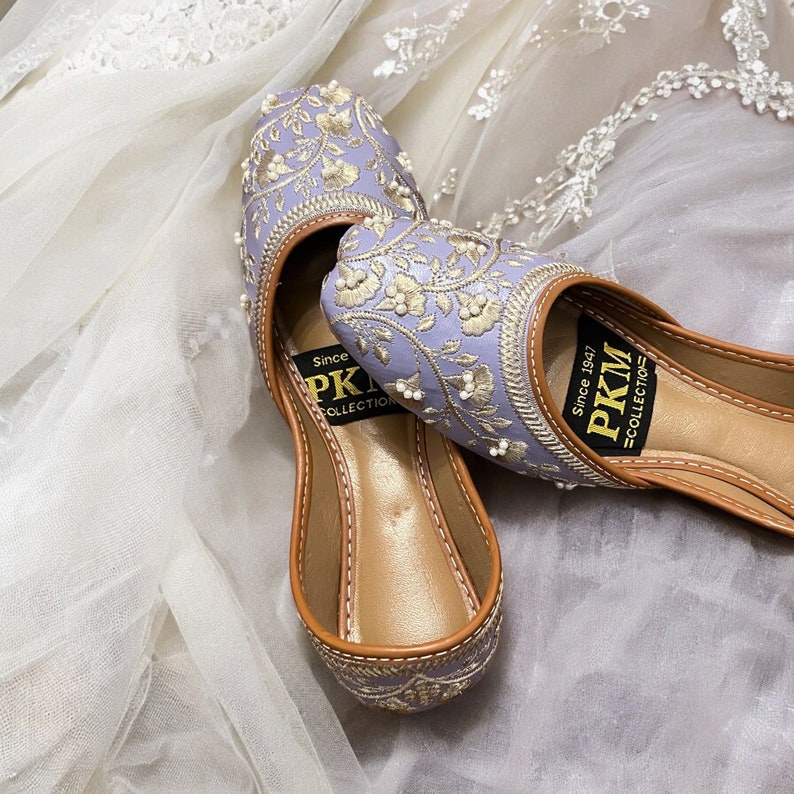 K&ouml;nnte beinhalten: Ein Paar flache, bestickte Schuhe in Lavendel mit goldenen F&auml;den und Perlenbesatz. Die Schuhe haben eine braune Ledersohle und sind mit "PKM Collection" und dem Text "Since 1947" beschriftet.
