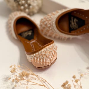 May include: A pair of peach-colored flat shoes with intricate pearl embellishments. The shoes have a pointed toe and a low heel. The embellishments are arranged in a geometric pattern and include a fringe of pearls around the ankle.