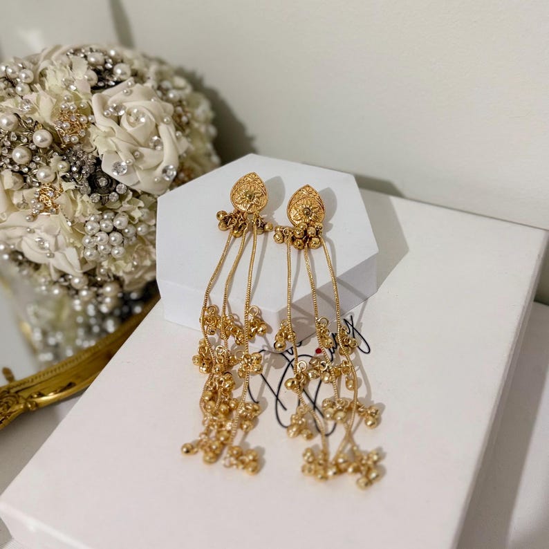 May include: A pair of gold-colored dangle earrings. Each earring features a floral design at the top, with multiple strands of delicate chains adorned with small, round beads. The earrings are displayed on a white surface.