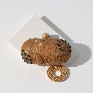 May include: A gold-toned oval clutch bag featuring intricate embroidery of peacocks and paisley patterns. The surface is embellished with small white beads and dark green crystals. A detachable gold beaded chain strap is coiled underneath.