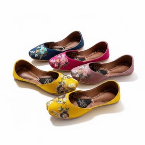 May include: Five flat slip-on shoes in blue, magenta, mauve, and two yellow pairs. Each shoe features intricate floral embroidery on the toe area, a rounded closed toe, and a brown interior lining with a black branded insole insert.