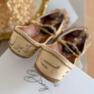 Puede incluir: Un par de zapatos bordados beige con adornos dorados. Los zapatos tienen una punta afilada y una correa en el tobillo. Se puede ver texto en un idioma extranjero en el lateral. Los zapatos est&aacute;n sobre una superficie blanca.