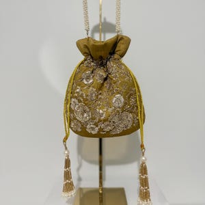 May include: A gold-colored drawstring bag with intricate floral embroidery. The bag features a pearl beaded strap and gold tassels. The bag is displayed on a gold stand against a white background. The bag's design is ornate and elegant.