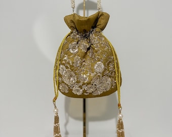以下が含まれることがあります： 金色の巾着バッグ。繊細な花の刺繍が施されています。パールビーズのストラップとゴールドのタッセルが特徴です。白い背景に、ゴールドのスタンドに展示されています。バッグのデザインは華やかでエレガントです。
