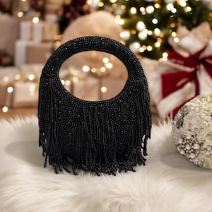 May include: A black beaded handbag with a circular handle and fringe detail. The bag is covered in small, round beads and has a soft, textured fringe hanging from the bottom. The bag is sitting on a white, fluffy surface.