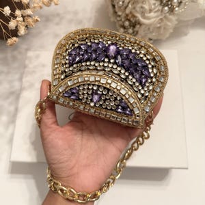 May include: A gold-toned, embellished clutch with a chain strap. The clutch is encrusted with purple and clear gemstones, arranged in a decorative pattern. The bag is held by a person, showcasing the gold chain strap and the intricate details of the design.