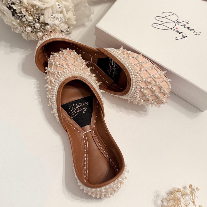 May include: A pair of pink bridal shoes with pearl embellishments. The shoes have a brown leather sole and are decorated with a delicate pearl pattern. The shoes are in a white box with the text "Dulhans Diary" on it.