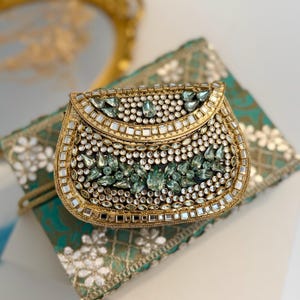 Op de afbeelding: Een gouden clutch met een afgeronde bovenkant en een klep sluiting. De clutch is versierd met verschillende heldere en groene strass steentjes.