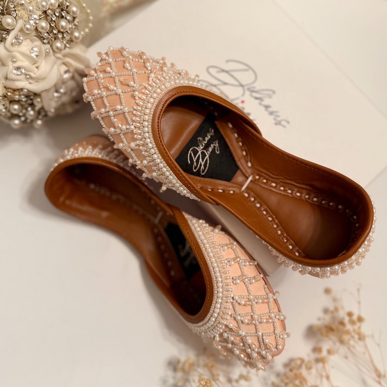 May include: A pair of pink and white embellished flat shoes with a brown leather insole. The shoes have a delicate pearl and bead design and the text "Delhish Diary" is visible on the insole.