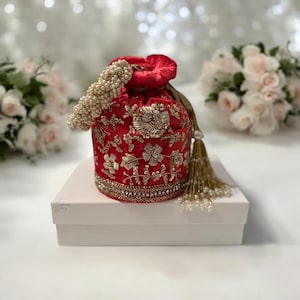 Może przedstawiać: Czerwona aksamitna torebka ze sznurkiem do ściągania, z haftem złotym i perłowymi akcentami. Torebka leży na białym pudełku z nadrukiem "Divine Joy".