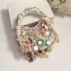 Può includere: Una borsetta bianca con perline, di forma curva e con un manico corto. La borsa è adornata con piccoli specchi, accenti colorati e perline pendenti a forma di perla. Il design incorpora vari colori, tra cui rosso, verde e arancione.