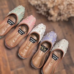 K&ouml;nnte beinhalten: F&uuml;nf Paar bestickte flache Schuhe in verschiedenen Farben, darunter Gr&uuml;n, Rosa, Wei&szlig;, Lila und Silber. Die Schuhe haben eine braune Ledersohle und sind mit goldenen Stickereien verziert. Die Schuhe sind mit "Dulhans Diary" und "PKM Collections since 1847" beschriftet.