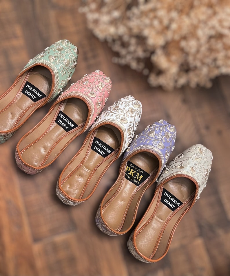 K&ouml;nnte beinhalten: F&uuml;nf Paar bestickte flache Schuhe in verschiedenen Farben, darunter Gr&uuml;n, Rosa, Wei&szlig;, Lila und Silber. Die Schuhe haben eine braune Ledersohle und sind mit goldenen Stickereien verziert. Die Schuhe sind mit "Dulhans Diary" und "PKM Collections since 1847" beschriftet.