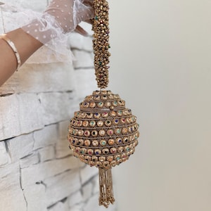 May include: A gold-colored, beaded handbag with a spherical shape. The bag is adorned with colorful gemstones and a beaded handle. A tassel hangs from the bottom. The bag is held up by a person wearing a white glove.