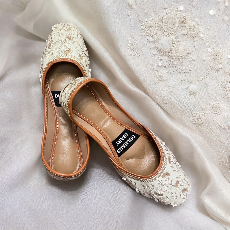 K&ouml;nnte beinhalten: Ein Paar wei&szlig;e, bestickte flache Schuhe mit braunen Ledersohlen. Die Schuhe haben Perlenverzierungen und der Text "Dulhans Diary" ist auf der Sohle aufgedruckt.