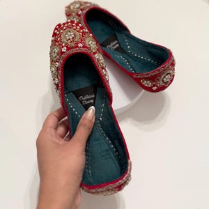K&ouml;nnte beinhalten: Ein Paar rote Samtschuhe mit goldenen und perlmuttartigen Verzierungen. Die Schuhe haben ein t&uuml;rkisfarbenes Futter und der Text "Dulhans Diary" ist auf der Innenseite eines Schuhs aufgedruckt.