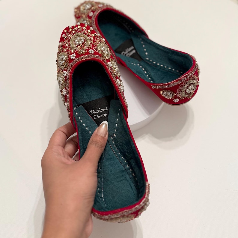 K&ouml;nnte beinhalten: Ein Paar rote Samtschuhe mit goldenen und perlmuttartigen Verzierungen. Die Schuhe haben ein t&uuml;rkisfarbenes Futter und der Text "Dulhans Diary" ist auf der Innenseite eines Schuhs aufgedruckt.