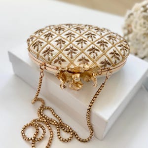 May include: An ivory-colored, oval-shaped clutch with a gold chain strap. The clutch features a quilted pattern with gold floral embellishments. A gold flower clasp with a pearl center adorns the front. The bag is resting on a white surface.