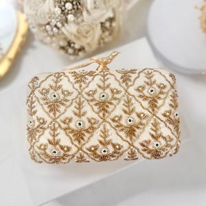 Könnte beinhalten: Eine cremefarbene, rechteckige Clutch mit aufwendigen goldenen Perlenstickereien in einem sich wiederholenden floralen Gittermuster. Die Tasche ist mit einem kleinen, goldfarbenen Verschluss in Vogelform versehen, der mit kleinen Kristallen besetzt ist.