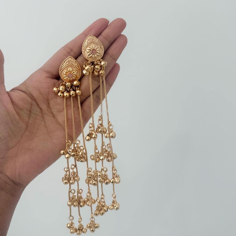 May include: Gold-colored earrings with a teardrop-shaped top featuring floral and textured designs. The earrings have multiple dangling chains with small, round beads. The earrings are held against a plain white background.