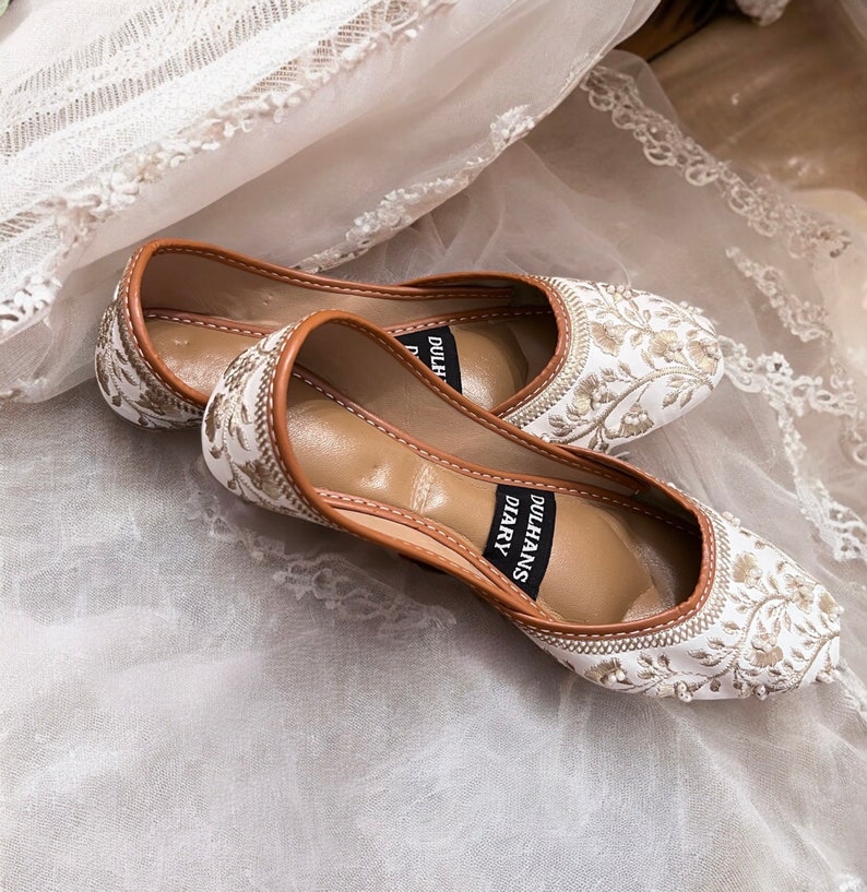 K&ouml;nnte beinhalten: Ein Paar wei&szlig;e, bestickte flache Schuhe mit braunem Lederbesatz. Die Schuhe haben ein Blumenmuster und der Text "Dulhans Diary" ist auf der Seite aufgedruckt.