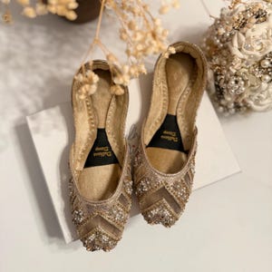 May include: A pair of gold-colored, embellished flats with intricate beadwork and a chevron pattern on the toe. The shoes have a soft, cushioned insole and a delicate, feminine design. The label "Dulhans Diary" is visible on the insole.