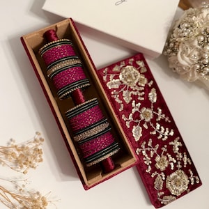 May include: A set of magenta bangles with gold and black accents, displayed in a rectangular, velvet-lined box. The box is open, revealing the stacked bangles. The lid of the box is also visible, featuring gold floral embroidery on a red velvet background.