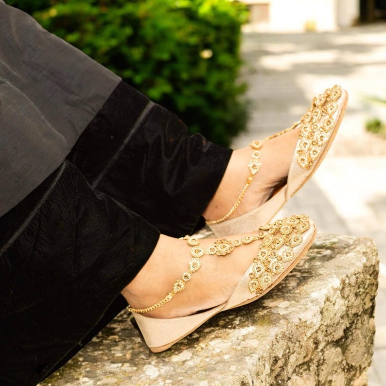 Puede incluir: Primer plano de sandalias doradas adornadas, con tac&oacute;n de cu&ntilde;a. Las sandalias presentan intrincados dise&ntilde;os florales y se combinan con una cadena de tobillo dorada. La persona lleva pantalones negros.