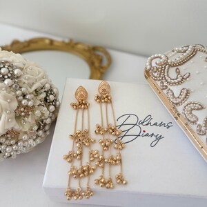 May include: Gold-toned dangle earrings with intricate detailing and cascading beaded strands. The earrings rest on a white box with the text "Dulhans Diary". A white clutch with pearl and gold embellishments is also visible.