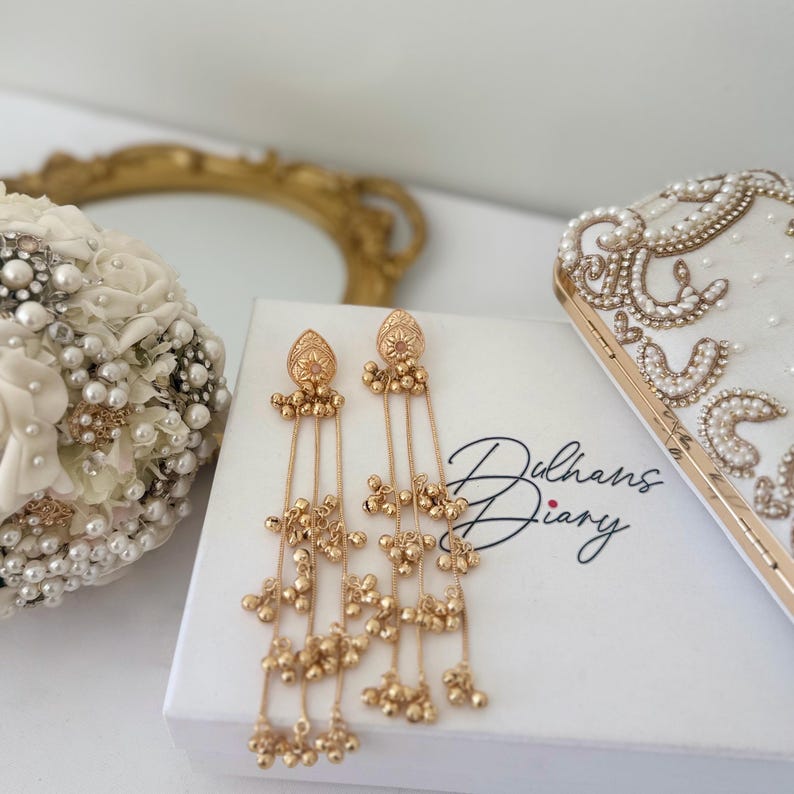 May include: Gold-toned dangle earrings with intricate detailing and cascading beaded strands. The earrings rest on a white box with the text "Dulhans Diary". A white clutch with pearl and gold embellishments is also visible.