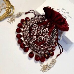 May include: A maroon velvet drawstring bag with gold and white embroidery and pearl accents. The bag has a tassel with white pearls and a matching tassel with maroon pom-poms.