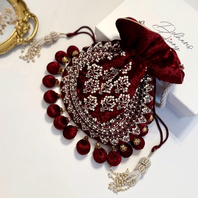 May include: A maroon velvet drawstring bag with gold and white embroidery and pearl accents. The bag has a tassel with white pearls and a matching tassel with maroon pom-poms.