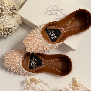 May include: A pair of peach-colored flat shoes with intricate beading and a brown leather sole. The shoes are decorated with a grid pattern and have the text "Delhvi Diary" on the sole.