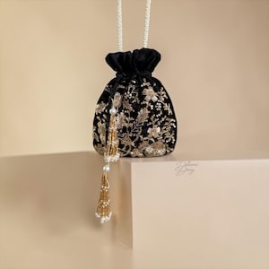 May include: Black velvet pouch bag with gold floral embroidery and a beaded tassel. The bag has a white beaded handle and is sitting on a white surface. The bag is a drawstring style.