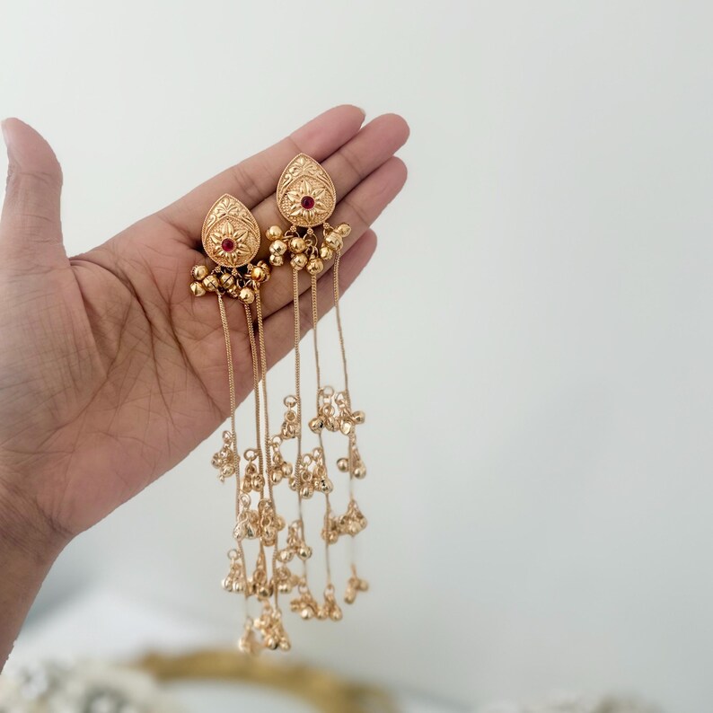 May include: Gold-toned dangle earrings with a teardrop-shaped top, featuring a small red gem and intricate floral detailing. Delicate chains cascade down, adorned with small, dangling gold beads. The earrings are designed for pierced ears.