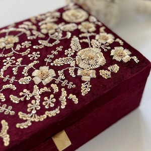 May include: A burgundy velvet box with intricate gold floral embroidery. The embroidery features detailed flowers, leaves, and delicate embellishments. The box has a rectangular shape and a gold-colored clasp.
