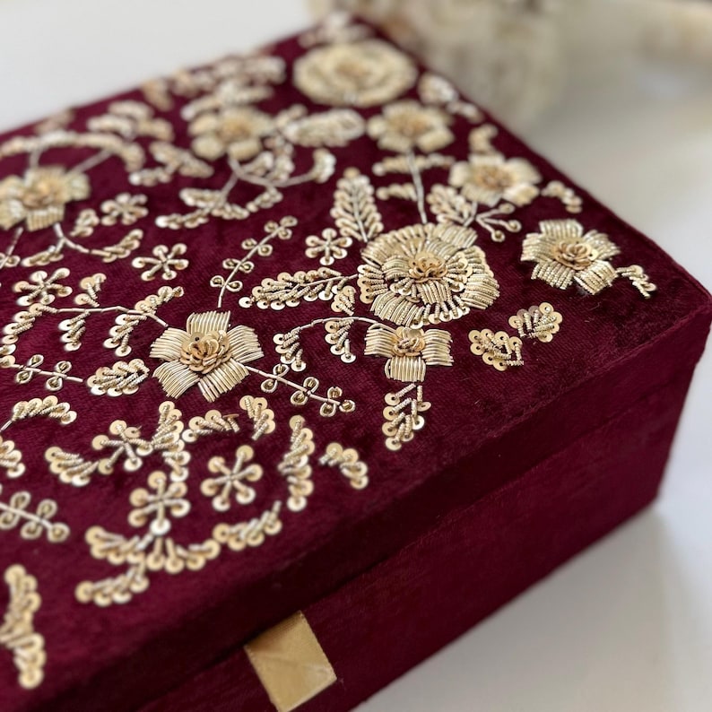 May include: A burgundy velvet box with intricate gold floral embroidery. The embroidery features detailed flowers, leaves, and delicate embellishments. The box has a rectangular shape and a gold-colored clasp.