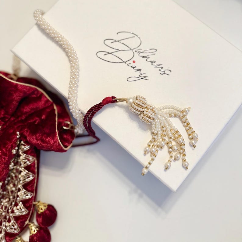 May include: A red velvet pouch with gold embroidery and a pearl tassel. The pouch is closed and sitting on a white surface. The pouch is part of a set that includes a white pearl necklace with a red cord.