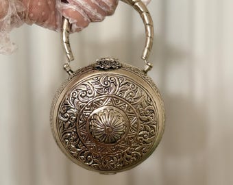 May include: A silver round purse with an ornate floral design and a silver handle.