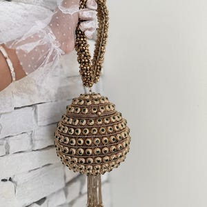 Pode incluir: Uma bolsa dourada com contas com um design redondo e em camadas. A bolsa apresenta pequenos detalhes verdes e uma borla. A alça também é feita de contas douradas. A bolsa é segurada contra uma parede de tijolos brancos.